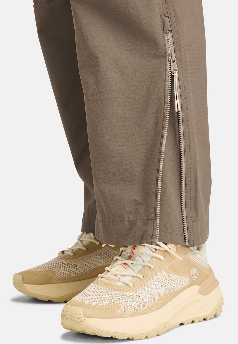Hellbraune Reißverschluss-Hosen kombiniert mit beigen Turnschuhen, die ein strukturiertes Netzobermaterial, eine Gummisohle und subtile Markenakzente aufweisen.