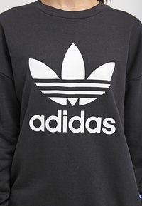 Černá mikina s kulatým výstřihem, která má velké bílé logo Adidas ve tvaru trojlístku a tři horizontální pruhy. Měkká textura, volný střih.