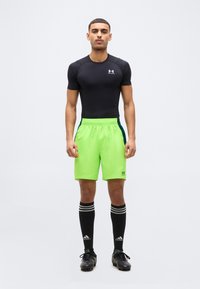 Αθλητής όρθιος, φοράει μαύρο ελαστικό μπλουζάκι, φωσφοριζέ πράσινο σορτς, μαύρες κάλτσες Adidas μέχρι το γόνατο και μαύρα παπούτσια με καρφιά, μπροστά σε απλό φόντο.