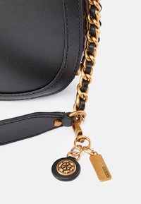 Černá kožená taška v rohu s zlatým řetízkovým popruhem a dvěma zlatými přívěsky, jedním kulatým s květinovým vzorem a jedním obdélníkovým s rytím 'GUESS'.