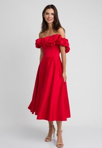 Czerwona sukienka bez ramiączek z falbanami, dopasowanym gorsetem i spódnicą w kształcie litery A. Gładki materiał o wyraźnej strukturze. Zestawiona z cielistymi szpilkami.