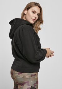 Felpa nera con maniche a sbuffo e cappuccio con lacci. Tessuto morbido con orlo a coste. Indossata con leggings a fantasia dai colori mimetici.