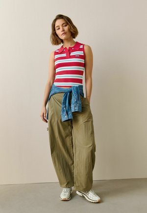 Młoda kobieta ubrana w czerwoną i białą pasiastą bluzkę bez rękawów, szerokie oliwkowe spodnie, białe trampki oraz niebieską dżinsową kurtkę zawiązaną w pasie.
