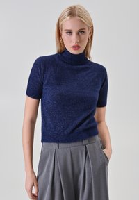 Donna con un maglione a polo blu navy a maniche corte e pantaloni grigi a vita alta con pieghe, con una mano in tasca, su uno sfondo neutro.