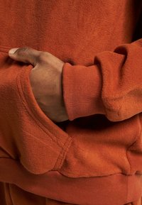 Oranžna majica s kapuco z mehkobo, flisasto teksturo, ki ima kenguru žep in rebrast detajl na manšetah. Poglej v bližini, roka resting v žepu.