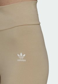 Nahaufnahme von beigen High-Waist Leggings mit dem weißen Adidas-Logo an der linken Hüfte vor einem hellen Hintergrund.