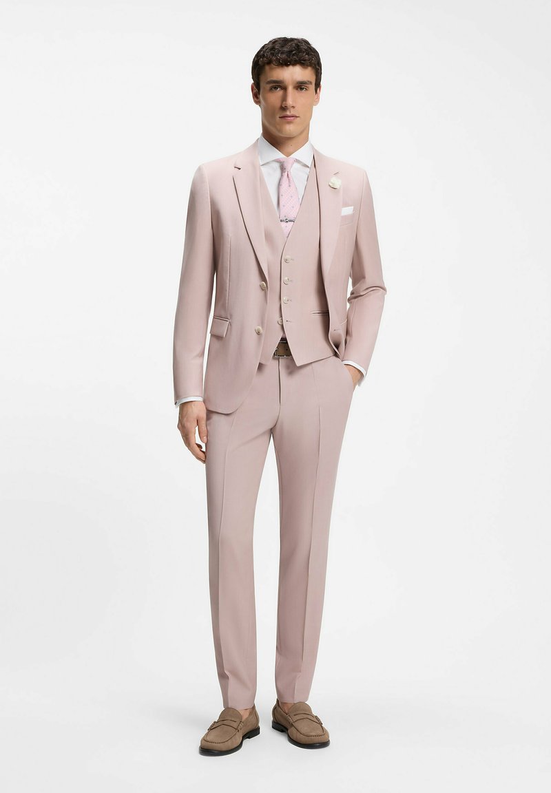 Homme portant un costume trois pièces rose clair, chemise blanche, cravate rose à motifs, mocassins beiges, debout devant un fond blanc uni.