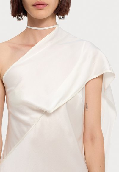 White silk asymmetrical top with draped texture, thin strap around the neck, and silver hardware earrings. Smooth finish and soft sheen.