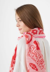 Écharpe rose avec des motifs graphiques rouges, y compris des cœurs et le texte "SUPER WOMAN." Fabriquée en tissu léger, elle tombe doucement sur un haut blanc.