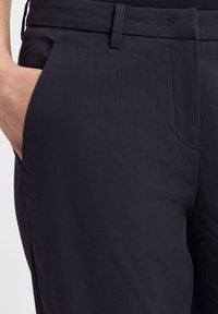 Dunkelblaue Hose mit glatter Textur, seitlichen Taschen und taillierter Passform. Enthält einen flachen Bund und dezente Nähte.