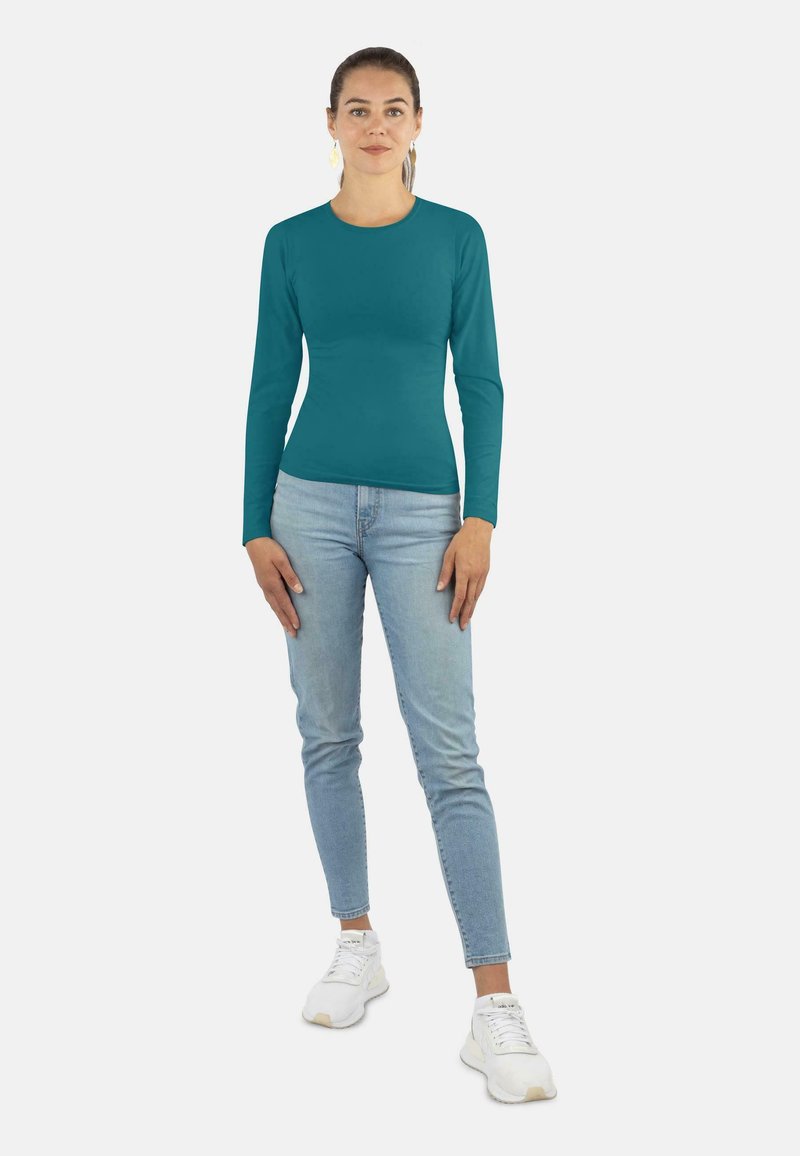 Teal Langarmshirt mit rundem Halsausschnitt, kombiniert mit hellblauen Jeans und weißen Sneakern. Einfaches, tailliertes Design, glatte Textur.