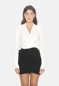 Blusa bianca a maniche lunghe con design avvolgente, abbinata a una mini gonna nera con arricciature e una texture liscia, dettaglio con bottoni dorati.