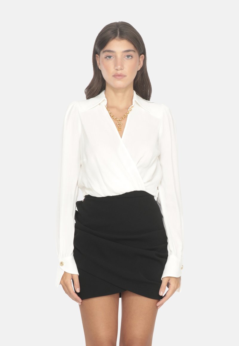 Blusa bianca a maniche lunghe con design avvolgente, abbinata a una mini gonna nera con arricciature e una texture liscia, dettaglio con bottoni dorati.