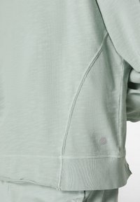 Sudadera de color verde claro, con una textura suave y un corte relajado, que presenta un detalle en la costura lateral y un pequeño logo en el dobladillo inferior.