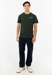 Man die tegen een witte achtergrond staat, draagt een donkergroen T-shirt met het logo "Protest", donkere jeans en witte sneakers.