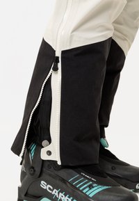 Schwarz-weiße Skihose mit geöffnetem Reißverschluss am unteren Bein, der den Skistiefel mit blauen Akzenten und Verschlussdetails freilegt.