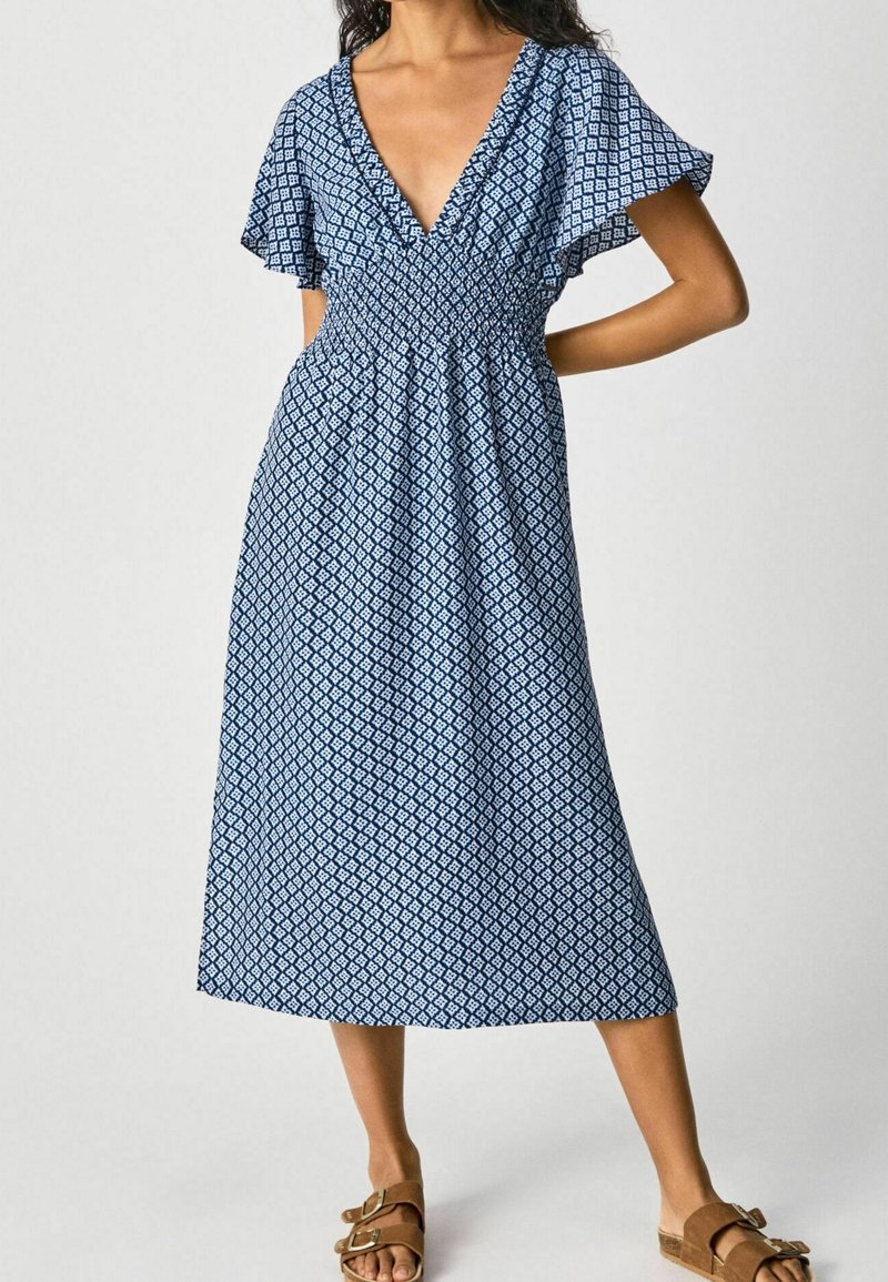 Femme portant une robe midi bleue à motifs avec un décolleté en V profond et des manches volantées, associée à des sandales à boucle couleur camel, debout devant un fond uni.
