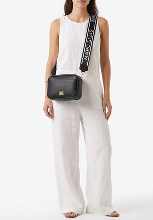 Donna che indossa una tuta bianca senza maniche e sandali, porta una borsa a tracolla in pelle nera con una tracolla larga etichettata "Marc Ellis."