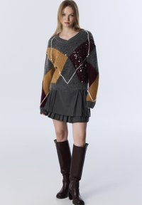 Strickpullover mit einem grau, gelb und burgunderfarbenen Rautenmuster, akzentuiert mit Pailletten; kombiniert mit einem dunkelgrauen, plissierten Rock und braunen kniehohen Stiefeln.