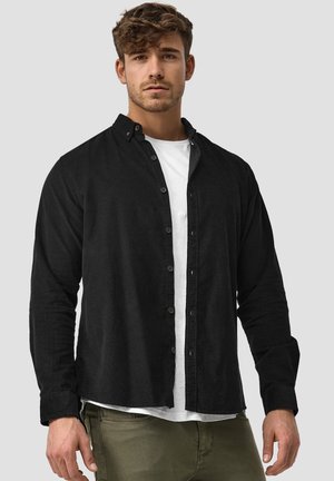 Zwarte button-up shirt gemaakt van een gestructureerde stof, met een kraag en lange mouwen, laag over een wit T-shirt. Olijfgroene broek zichtbaar.