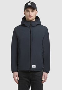 Mørkeblå jakke med frontlynlås, snorhætte, elastiske manchetter og et lille hvidt mærke ved kanten. Båret over mørke jeans.
