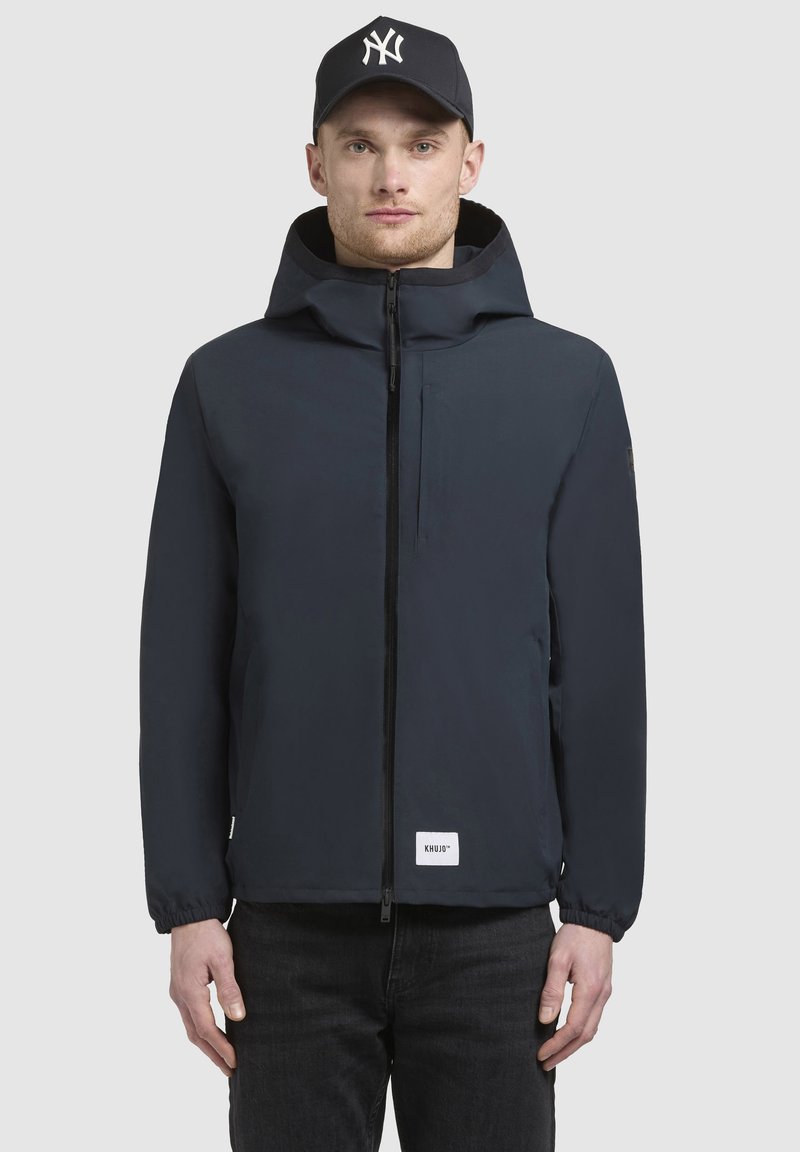 Mørkeblå jakke med frontlynlås, snorhætte, elastiske manchetter og et lille hvidt mærke ved kanten. Båret over mørke jeans.