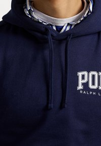 Námořnická modrá bavlněná mikina s kapucí a stahovacími šňůrkami, se bílým textem "Polo Ralph Lauren" v geometrickém písmu na hrudi a vrstveným límcem.