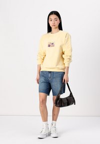 Gul crewneck sweatshirt med en framapplikation, kombinerat med denimshorts. Modellen bär vita högskurna sneakers och håller en svart handväska.
