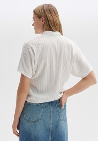 Weiße, strukturierte Bluse mit kurzen Ärmeln, die vertikale Streifen aufweist. Kombiniert mit einem Jeansrock, Rückansicht gezeigt.