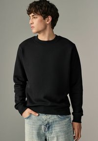 Sudadera negra de cuello redondo hecha de tela suave, con puños y dobladillo de canalé, combinada con jeans de mezclilla azul claro. Diseño liso, sin patrones.