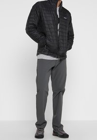 Svart quiltad jacka med hög krage, dragkedja framtill och sidofickor, kombinerad med grå byxor och mörka skor med röda detaljer.