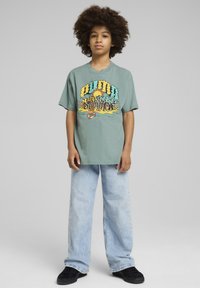 Camiseta de algodón verde menta con estampado gráfico multicolor y el texto "Verano Infinito". Combinada con unos vaqueros anchos de color azul claro y zapatos negros.