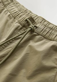 Shorts kaki avec une taille élastique et une fermeture à cordon, texture lisse et coutures décoratives le long de la ceinture.