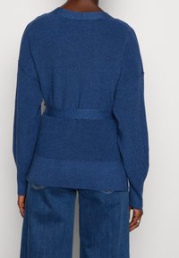 En person klädd i en blå stickad tröja med bred midja och lösa blå denimbyxor, sedd bakifrån mot en vit bakgrund.