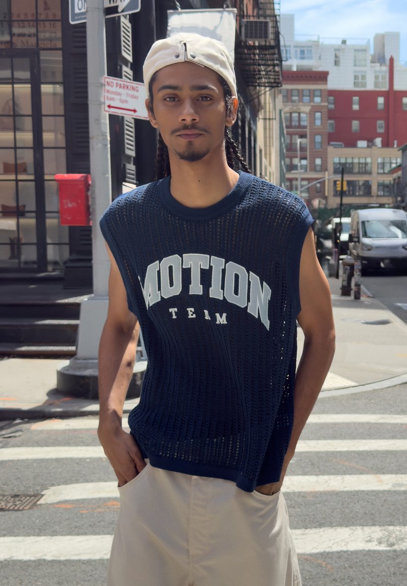 Jeune homme aux cheveux tressés portant une casquette beige à l'envers, un top en maille sans manches bleu marine avec la mention "MOTION TEAM" et un pantalon clair, debout sur un passage piéton en ville.