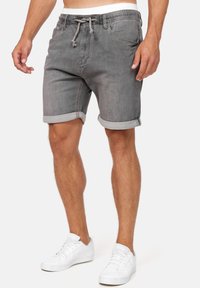 Shorts en denim gris avec des poignets roulés, une taille avec cordon de serrage ajustable et des poches avant. Portés avec des baskets blanches sur un fond neutre.
