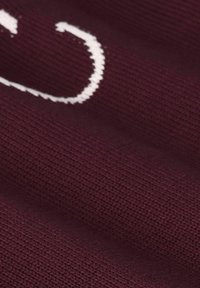 Close-up van kastanjebruine gebreide stof die de textuur laat zien en een deel van een witte gestikte patroon of letter op het oppervlak.