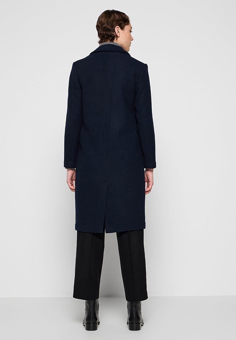 Marineblauer Wollmantel mit gerader Silhouette, Reverskragen und langen Ärmeln. Verfügt über einen Schlitz hinten und saubere Steppnähte. Kombiniert mit schwarzen Hosen.