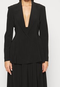Černý šitý blazer s hlubokým výstřihem ve tvaru V, strukturovanými ramenními vycpávkami a sklady v pase. Vyrobeno z hladkého, splývavého materiálu.
