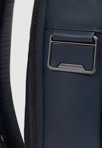 Bolso de cuero azul marino con textura suave, cuenta con una etiqueta de metal plateado y una cremallera en el borde. Los detalles incluyen costuras decorativas.