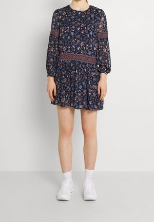 Personne portant une robe fleurie bleu foncé avec des bandes rouges à motifs sur les manches et la taille, des chaussettes blanches et des baskets blanches, debout devant un fond uni.