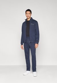 Μπλε φόρμα από μαλακό ύφασμα, με ζακέτα με φερμουάρ και ταιριαστό παντελόνι με στενή εφαρμογή, συνδυασμένη με λευκά αθλητικά παπούτσια.