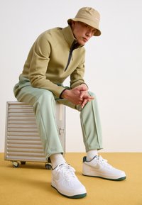 Zelená fleecová mikina na zip, světlezelené tepláky, bílé tenisky se zelenými akcenty a béžový klobouk typu bucket, sedící na stříbrném kufru.