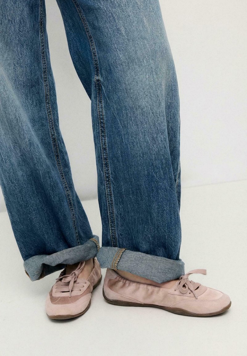 Personne portant un jean bleu retroussé et des ballerines lacées rose pâle, debout sur un sol blanc contre un mur uni.