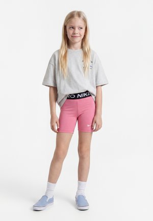 Jeune fille aux longs cheveux blonds portant un t-shirt gris oversize, un short Nike rose, des chaussettes blanches et des chaussures slip-on bleues, debout devant un fond blanc.
