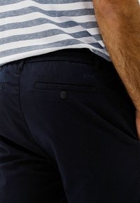 Marina shorts med en mjuk textur, med en bakficka med knapp och "Brax"-logotypen på midjebandet. Randerad skjorta delvis synlig.