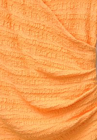 Texturovaná látka v jasně oranžové barvě, s vlnitým vzorem a skládajícími se záhyby, které zdůrazňují její lehkost a flexibilitu.