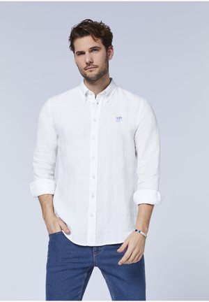 Weißes Leinen-Hemd mit langen Ärmeln, die bis zu den Ellenbogen aufgerollt sind, mit einem kleinen blauen Logo auf der Brust, kombiniert mit blauen Jeans.