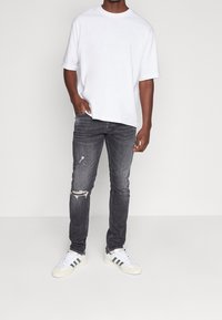 Vit t-shirt med kort ärm, slim-fit slitna grå jeans, och vita sneakers med svarta ränder, stående mot en ljusgrå bakgrund.