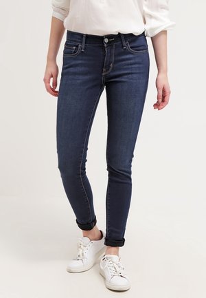 Jeans ajustados de mezclilla azul oscura con costuras claras, cintura de media altura y puños enrollados. Combinados con zapatillas blancas.